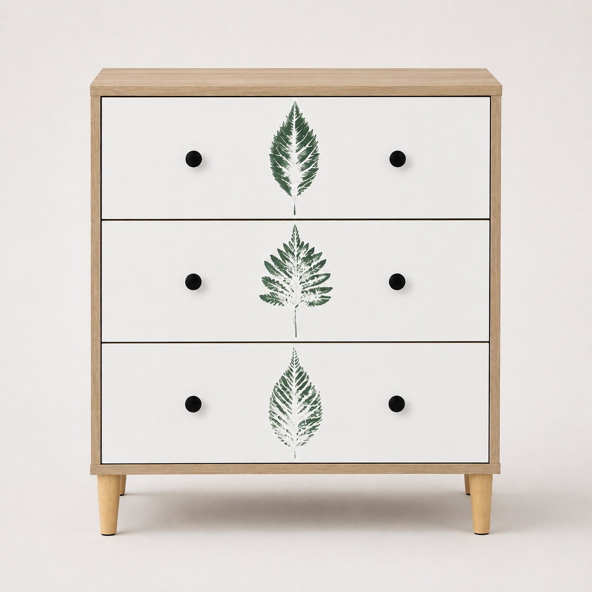 Commode 3 tiroirs design vue de face, finition bois clair, façades blanches à feuilles vertes et poignées noires.