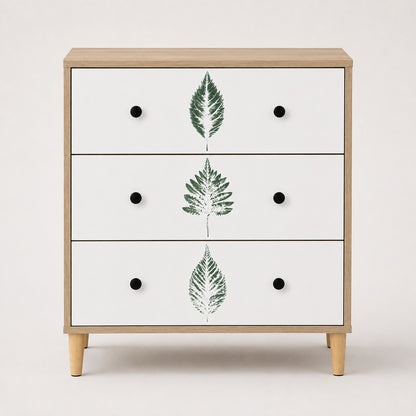Commode 3 tiroirs design vue de face, finition bois clair, façades blanches à feuilles vertes et poignées noires.