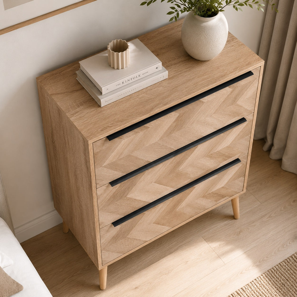 Vue en plongée d’une commode 3 tiroirs design en bois clair avec poignées noires et façades graphiques.