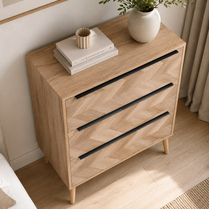 Vue en plongée d’une commode 3 tiroirs design en bois clair avec poignées noires et façades graphiques.